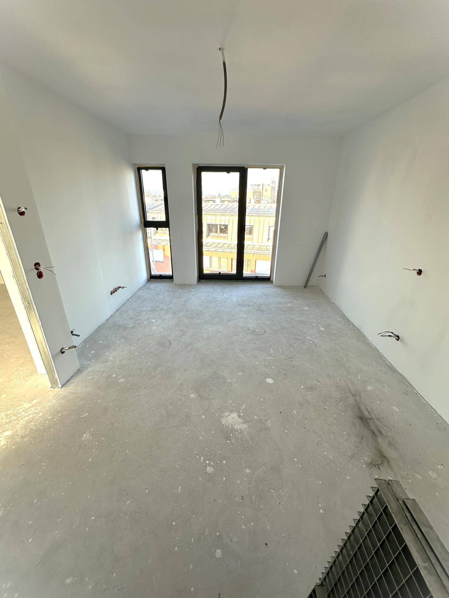 Unmöbliertes Zimmer im Rohbau mit zwei großen Fenstern, die viel Tageslicht in den Raum lassen.