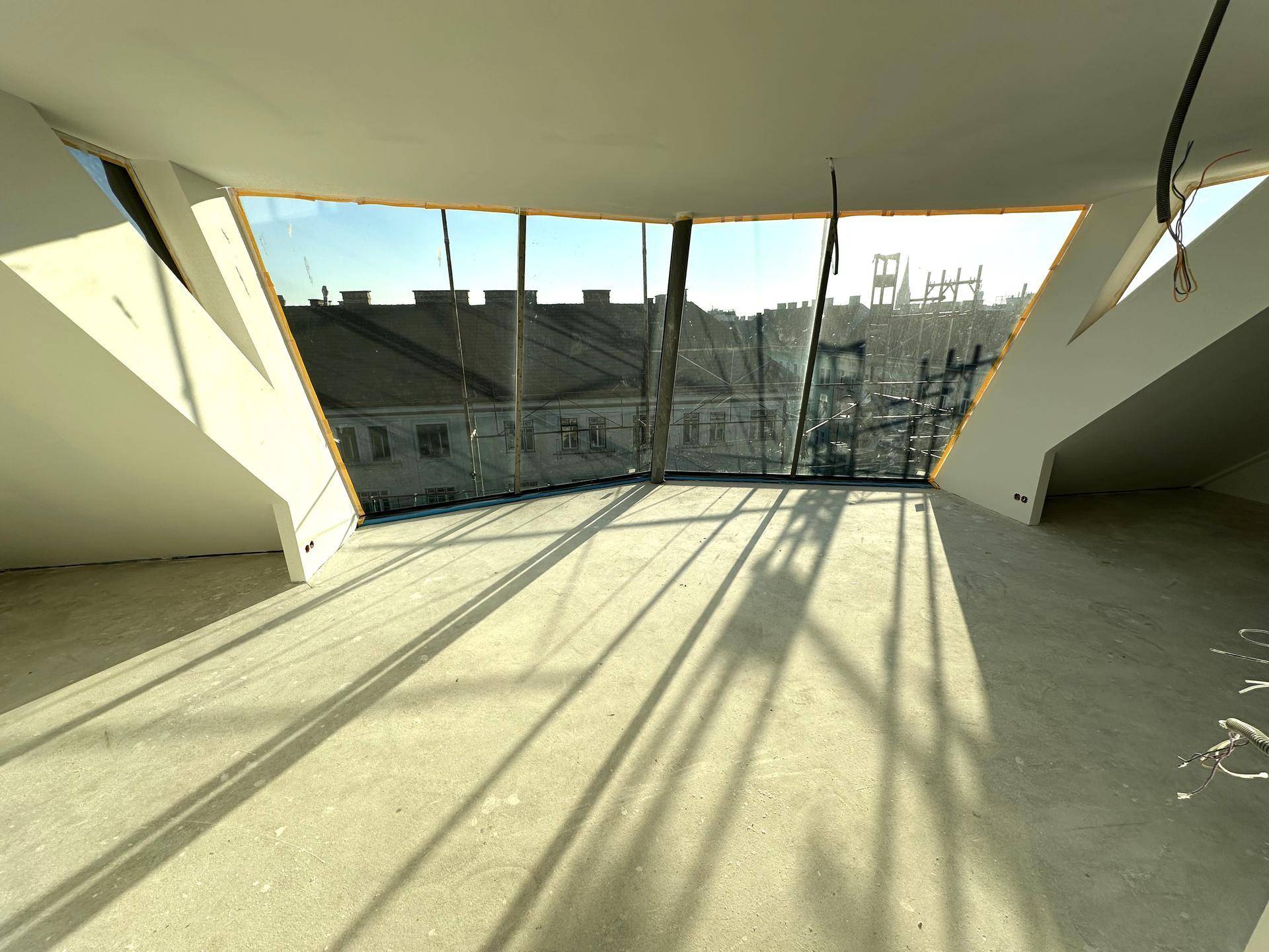 Heller Innenraum im Rohbau mit großen, schrägen Fenstern und Blick auf die Stadt, durchflutet von Sonnenlicht.