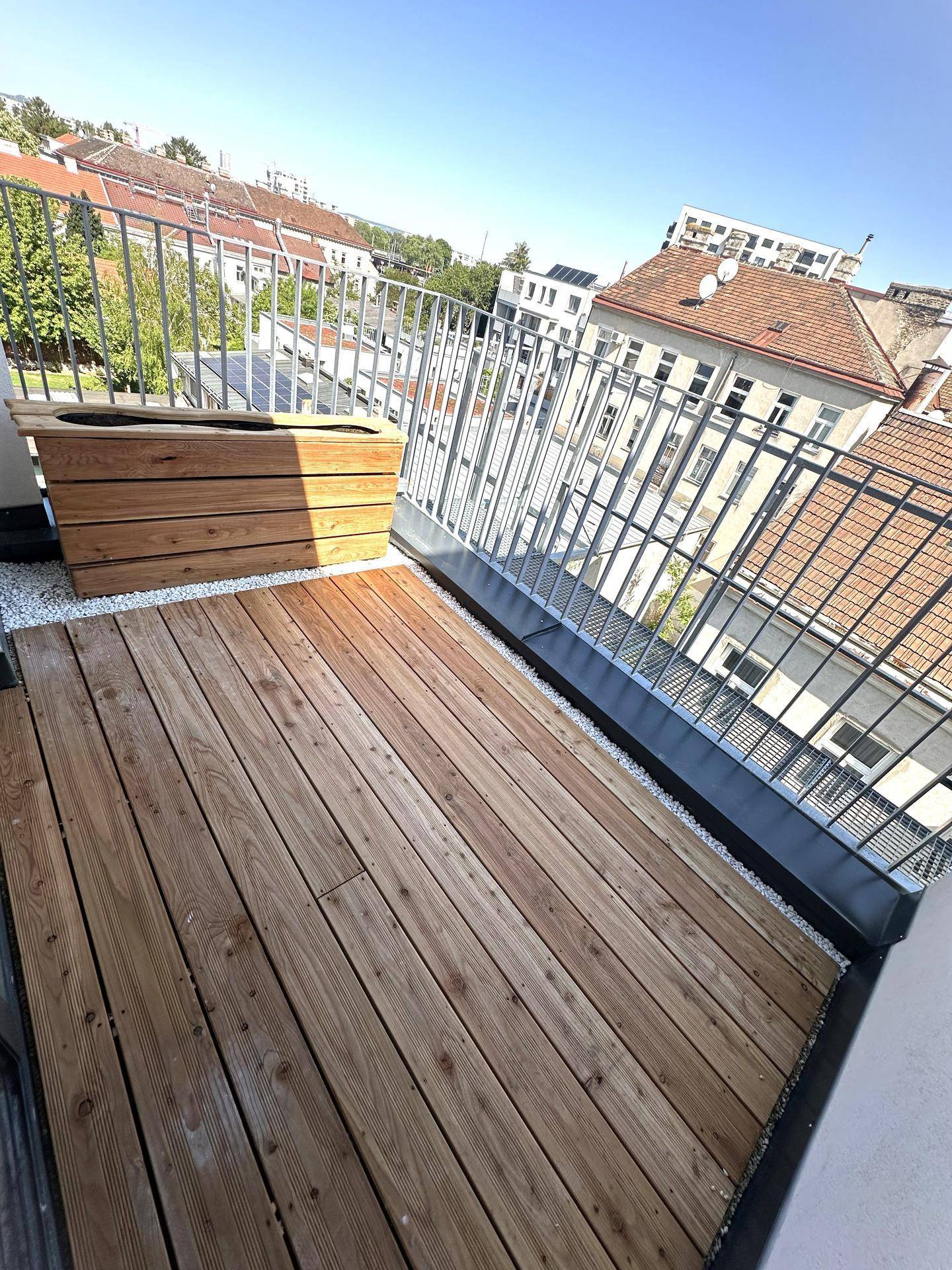 Kleiner Balkon mit Holzboden, Metallgeländer und einem Holzkasten für Pflanzen, sonnig und hell.