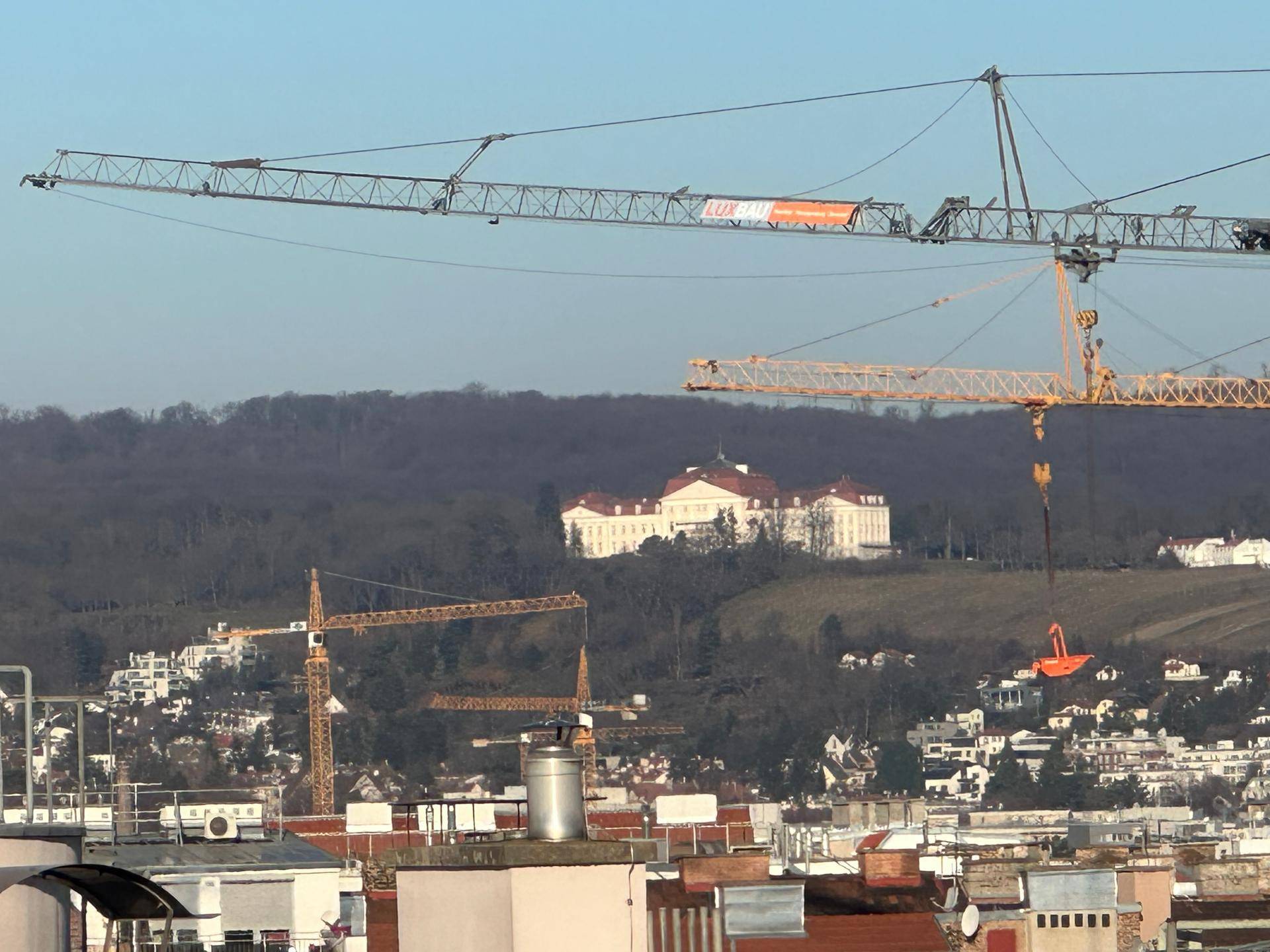 Panoramablick auf ein historisches Schloss auf einem Hügel mit Baukranen im Vordergrund und Stadtansicht.