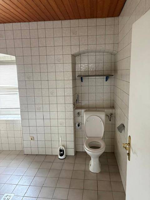 WC-Raum mit weißen Fliesen an Wänden und Boden sowie einer Holzdeckenverkleidung.