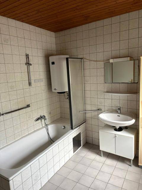 Badezimmer mit Badewanne, Waschbecken und Spiegelschrank, komplett gefliest und Holzdecke.