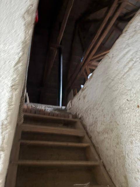 Steile Holztreppe führt zu einem Dachboden mit sichtbaren Holzbalken und Dämmung.