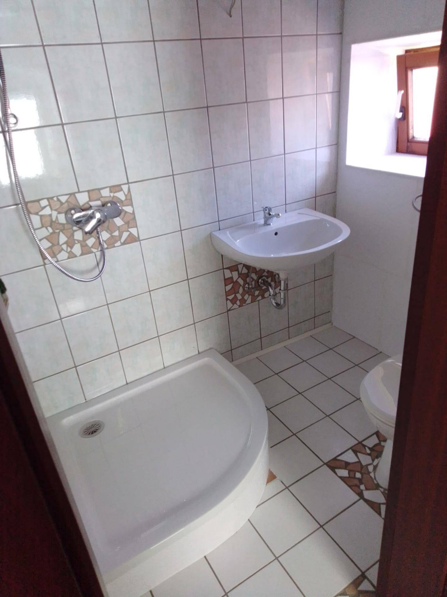 Helles Badezimmer mit weißen Fliesen, Duschkabine, Waschbecken und Toilette, ergänzt durch ein kleines Fenster.