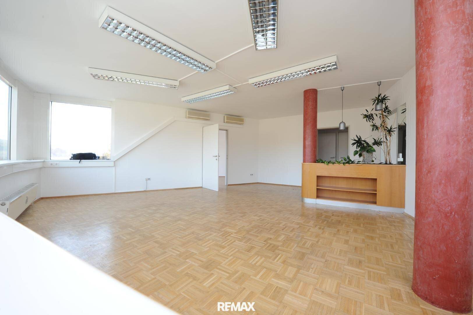 Geräumiges, leeres Büro mit Parkettboden, weißen Wänden und einem dekorativen roten Pfeiler.
