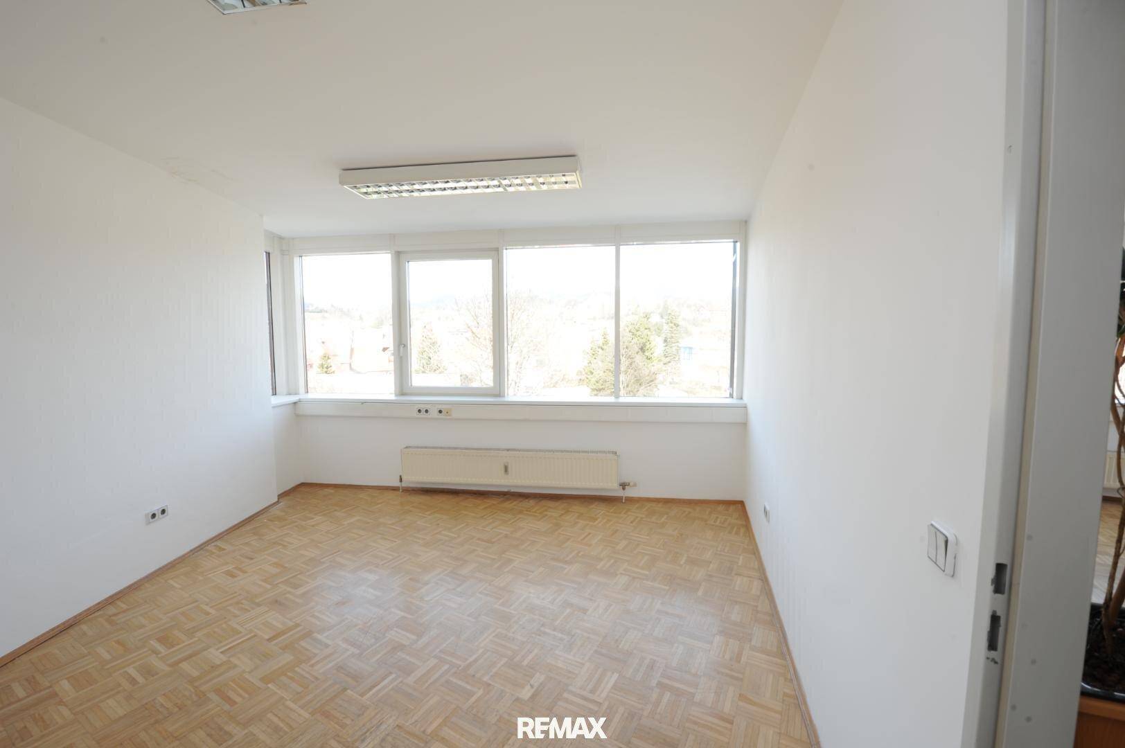 Leeres Büro mit hellen Wänden, Parkettboden und großen Fenstern für viel Tageslicht.