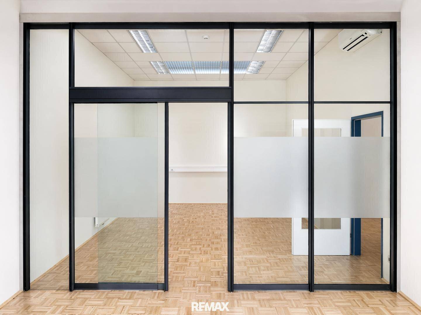 Modernes Büro mit Glaswänden und Schiebetür, ausgestattet mit Parkettboden und Deckenbeleuchtung.