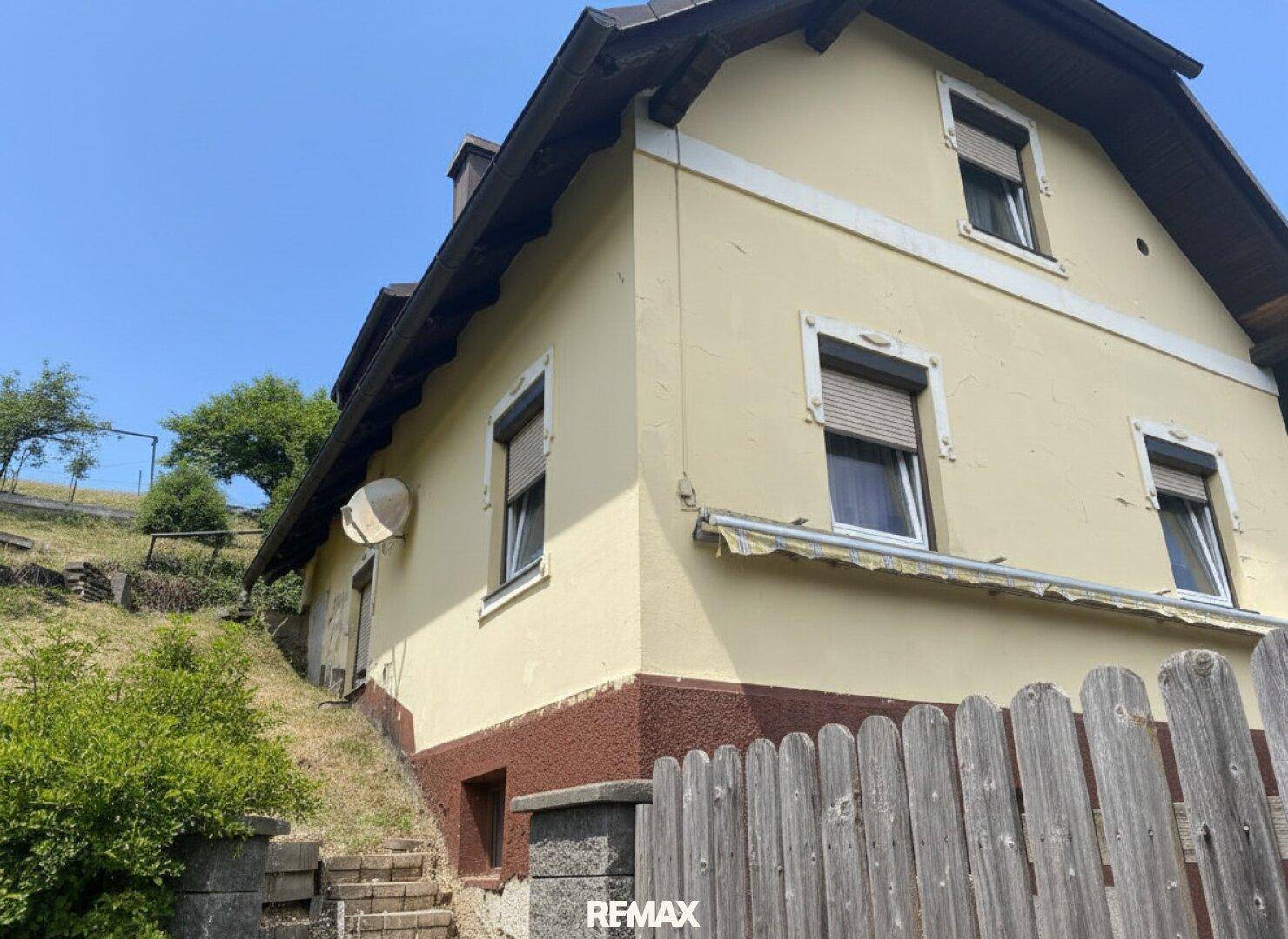 Seitenansicht des Hauses mit gelber Fassade, braunem Sockel und Fenstern mit Rollläden.