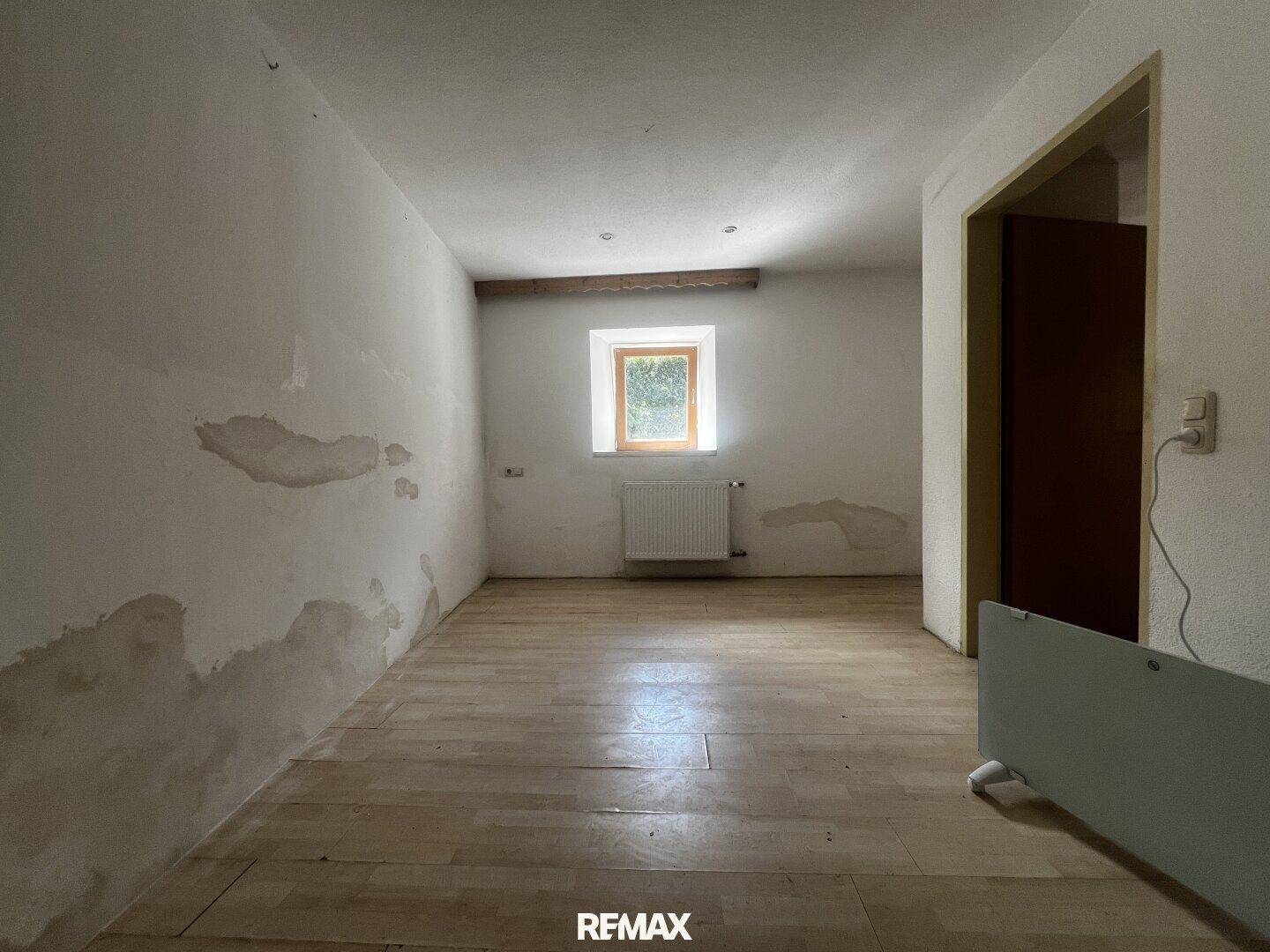 Helles Zimmer mit Holzfußboden, weißen Wänden und einem Heizkörper unter dem Fenster, mit Renovierungsbedarf.