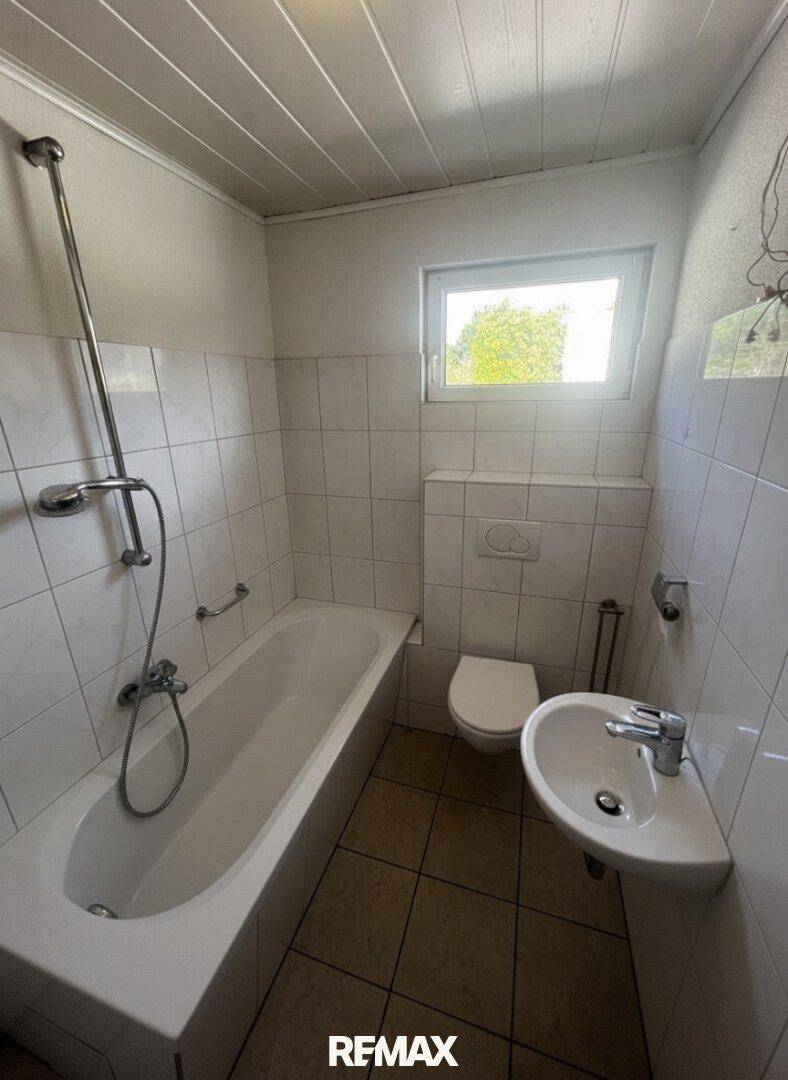 Badezimmer mit Badewanne, Toilette, Waschbecken und gefliesten Wänden, inklusive Fenster für Tageslicht.