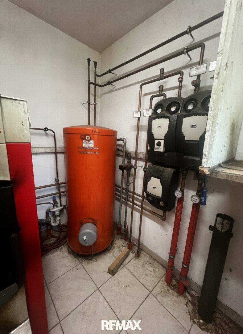 Technikraum mit großem Warmwasserspeicher, Rohrleitungen und Pumpen auf einem gefliesten Boden.