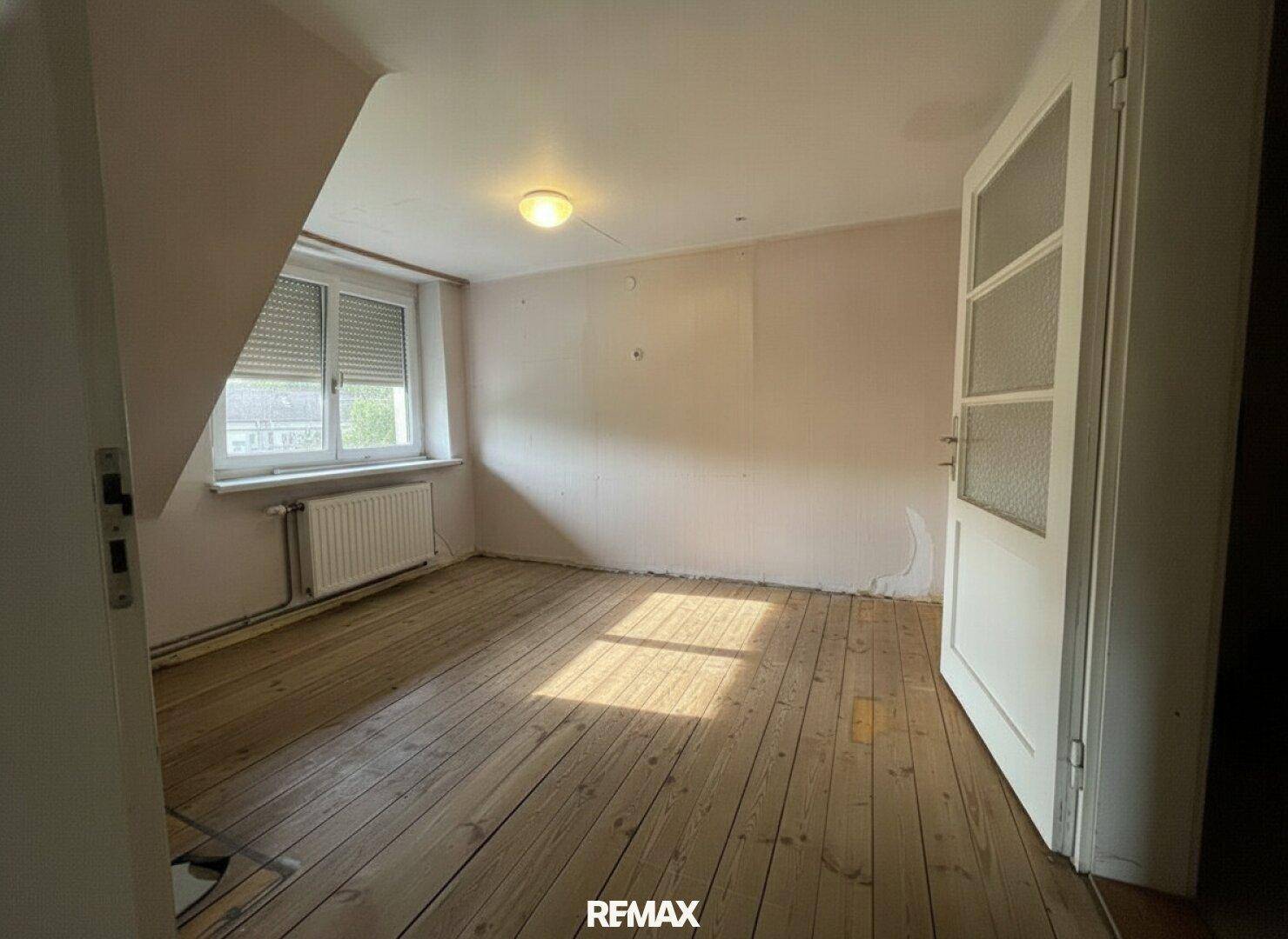 Schlafzimmer mit Holzdielenboden, schrägen Wänden und Fenster mit Rollladen, bereit zur Renovierung.