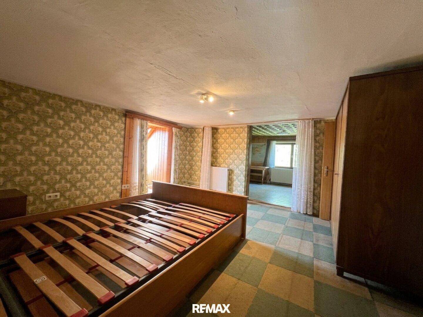Schlafzimmer mit gemusterter Tapete, Fliesenboden und Blick in einen angrenzenden Raum.