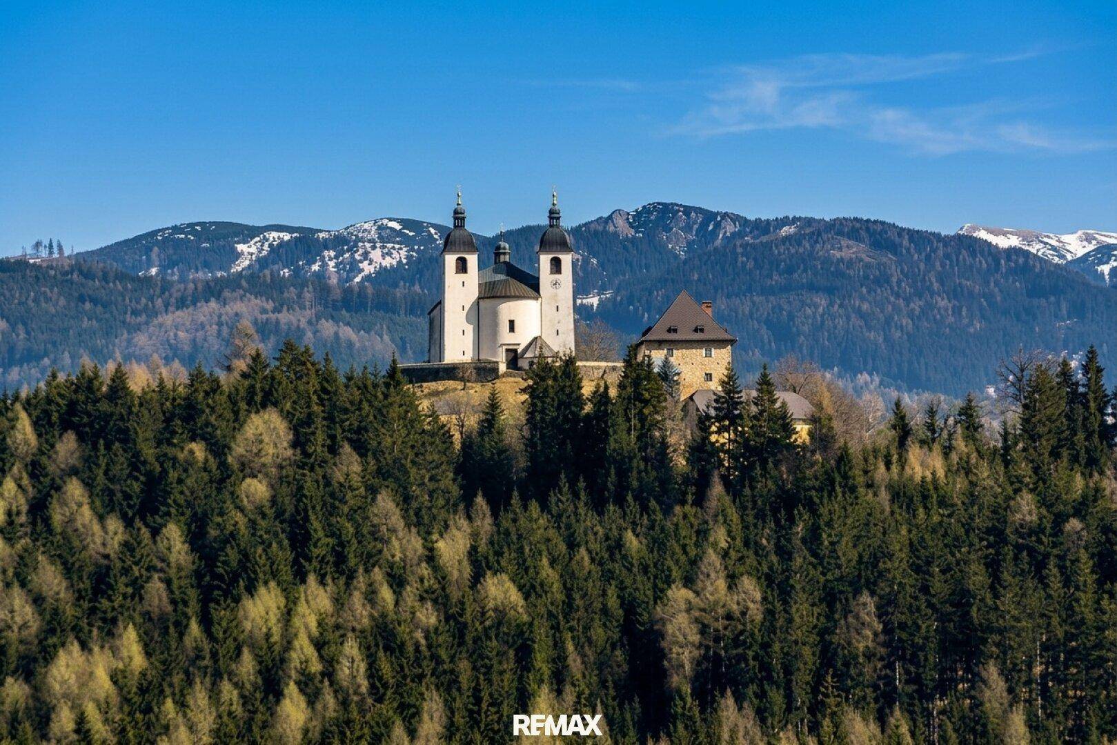 Malerische Kirche auf einem bewaldeten Hügel mit schneebedeckten Bergen im Hintergrund.