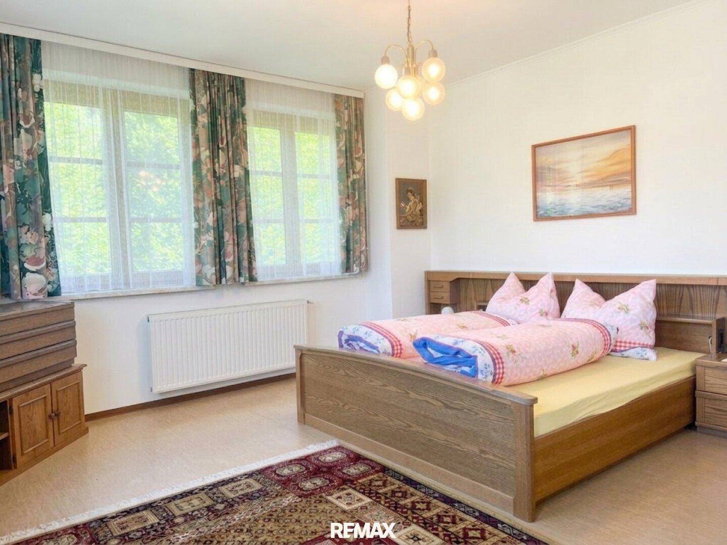 Helles Schlafzimmer mit Holzrahmenbett, rosa Bettwäsche und großen Fenstern mit floralen Vorhängen.