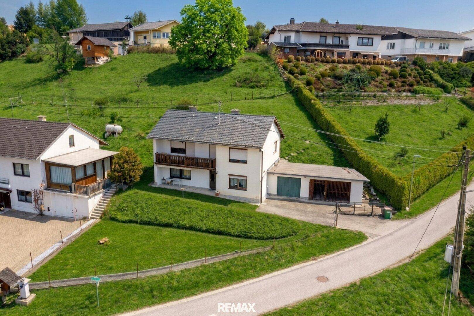 Luftaufnahme eines gepflegten Einfamilienhauses mit Garage, Balkon und großem Garten in grüner Hanglage.