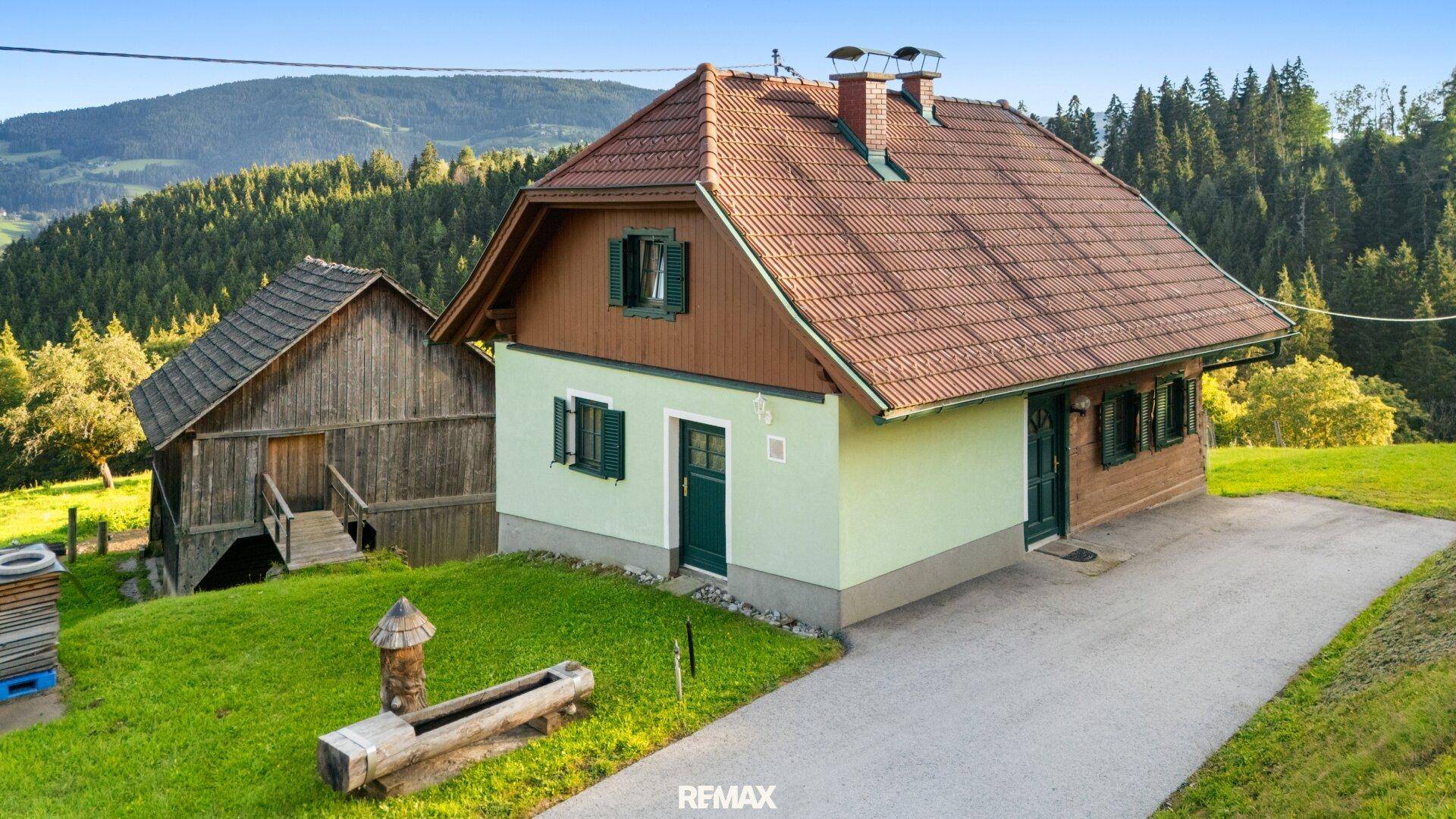 Traditionelles Landhaus mit rotem Ziegeldach, hellgrüner Fassade und grünen Fensterläden, umgeben von Natur.