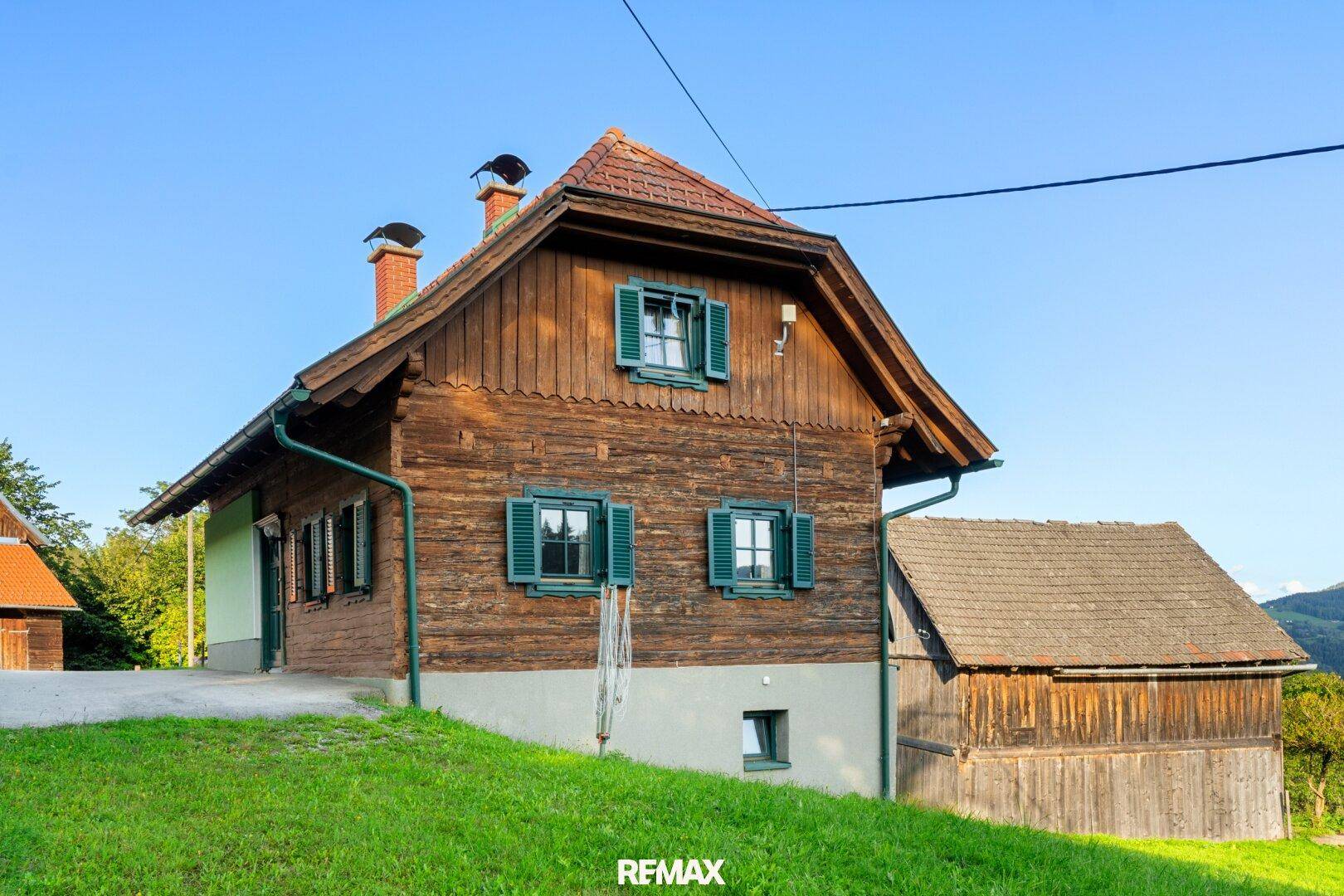 Seitenansicht des traditionellen Landhauses mit Holzverkleidung, grünen Fensterläden und ländlicher Umgebung.