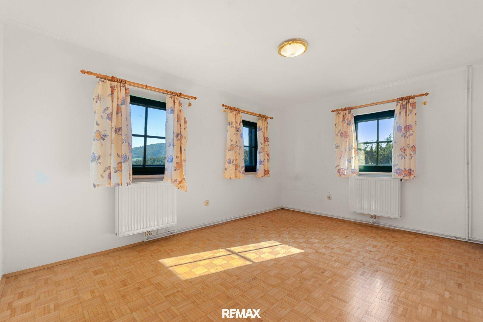Leeres Zimmer mit drei Fenstern, weißen Wänden und traditionellem Parkettboden, hell und sonnig.