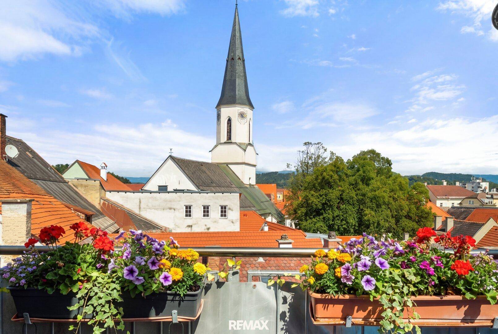 Balkon mit bunten Blumenkästen und malerischem Blick auf die Kirchturmspitze und die Stadt.