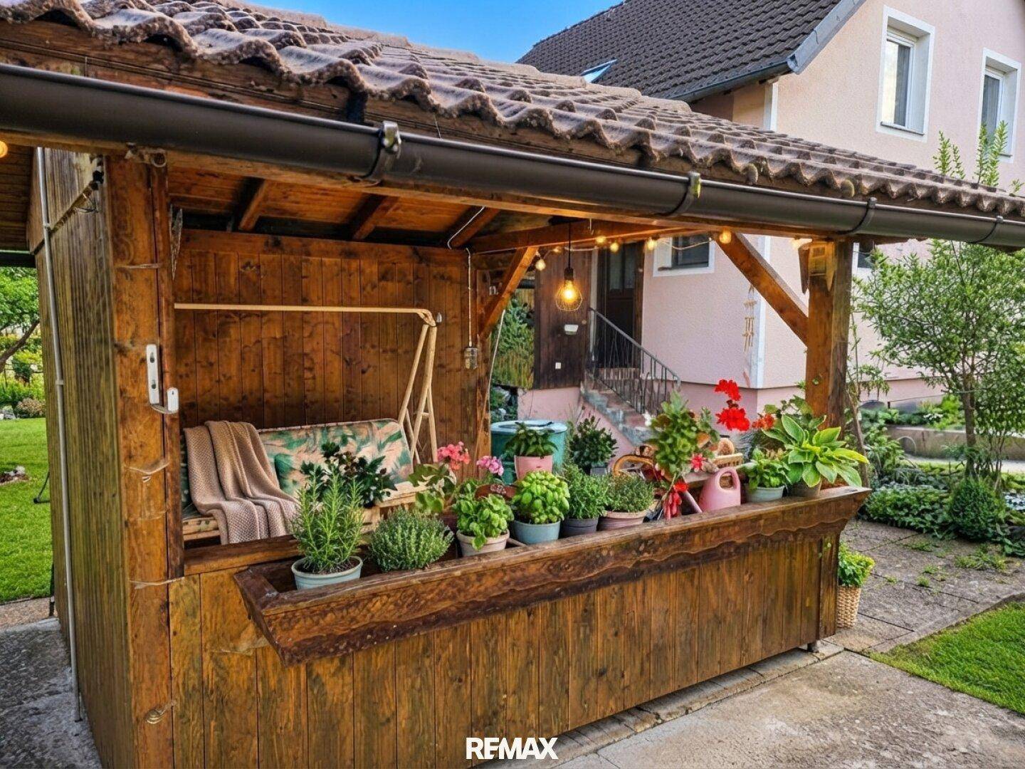 Überdachter Sitzbereich im Garten mit Schaukelbank und vielen Pflanzen in Töpfen.