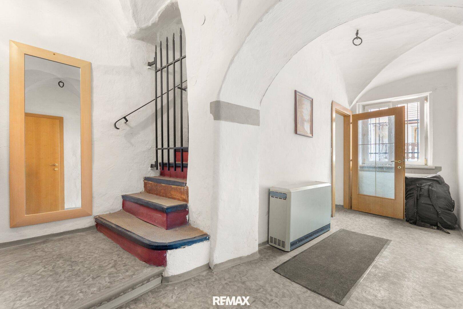 Heller Eingangsbereich mit Gewölbedecke, großem Spiegel und einer Treppe mit schmiedeeisernem Gitter.