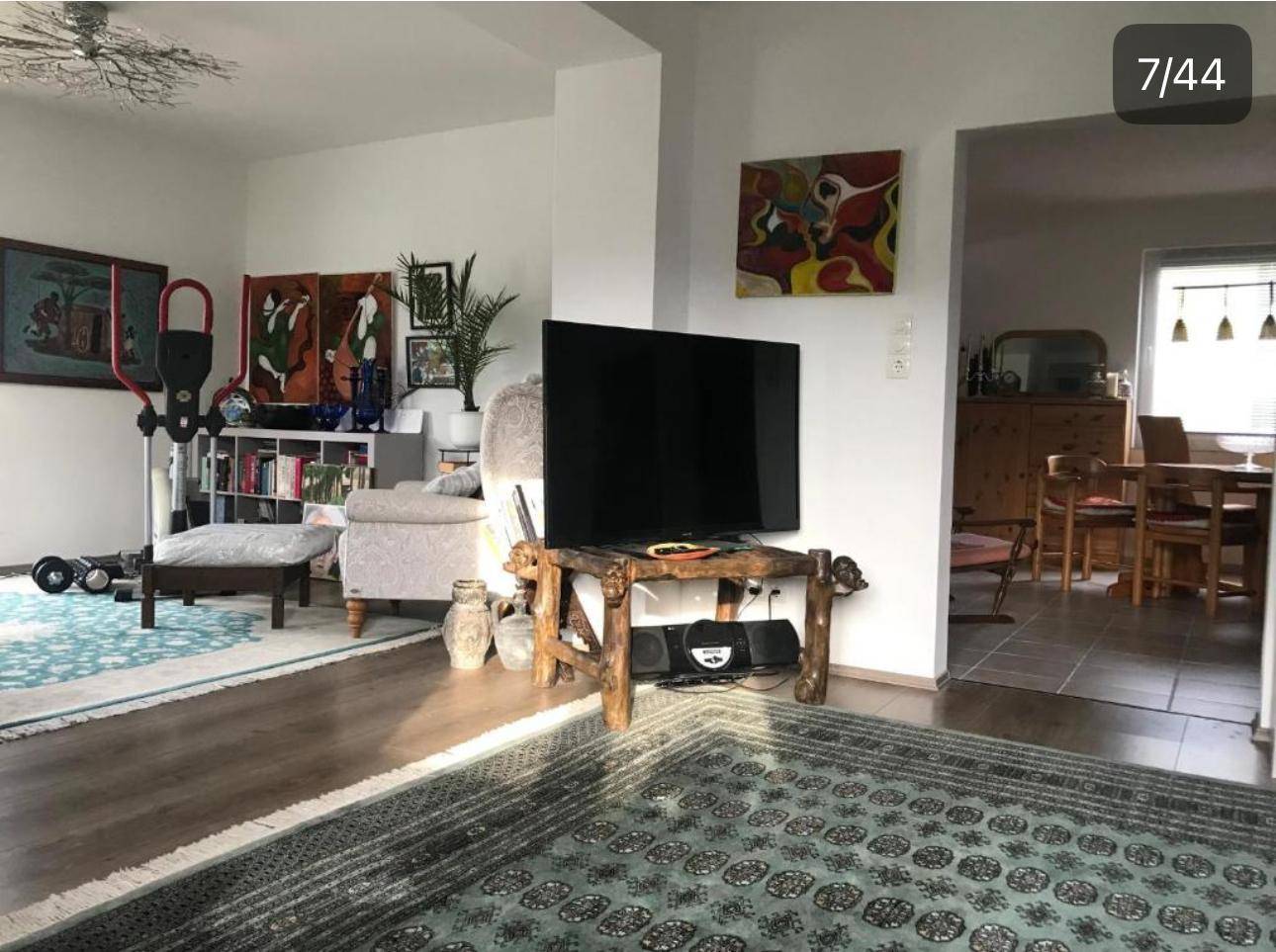 Wohnzimmer mit Fernseher, Trainingsgerät, Bücherregal und Übergang zum Essbereich.