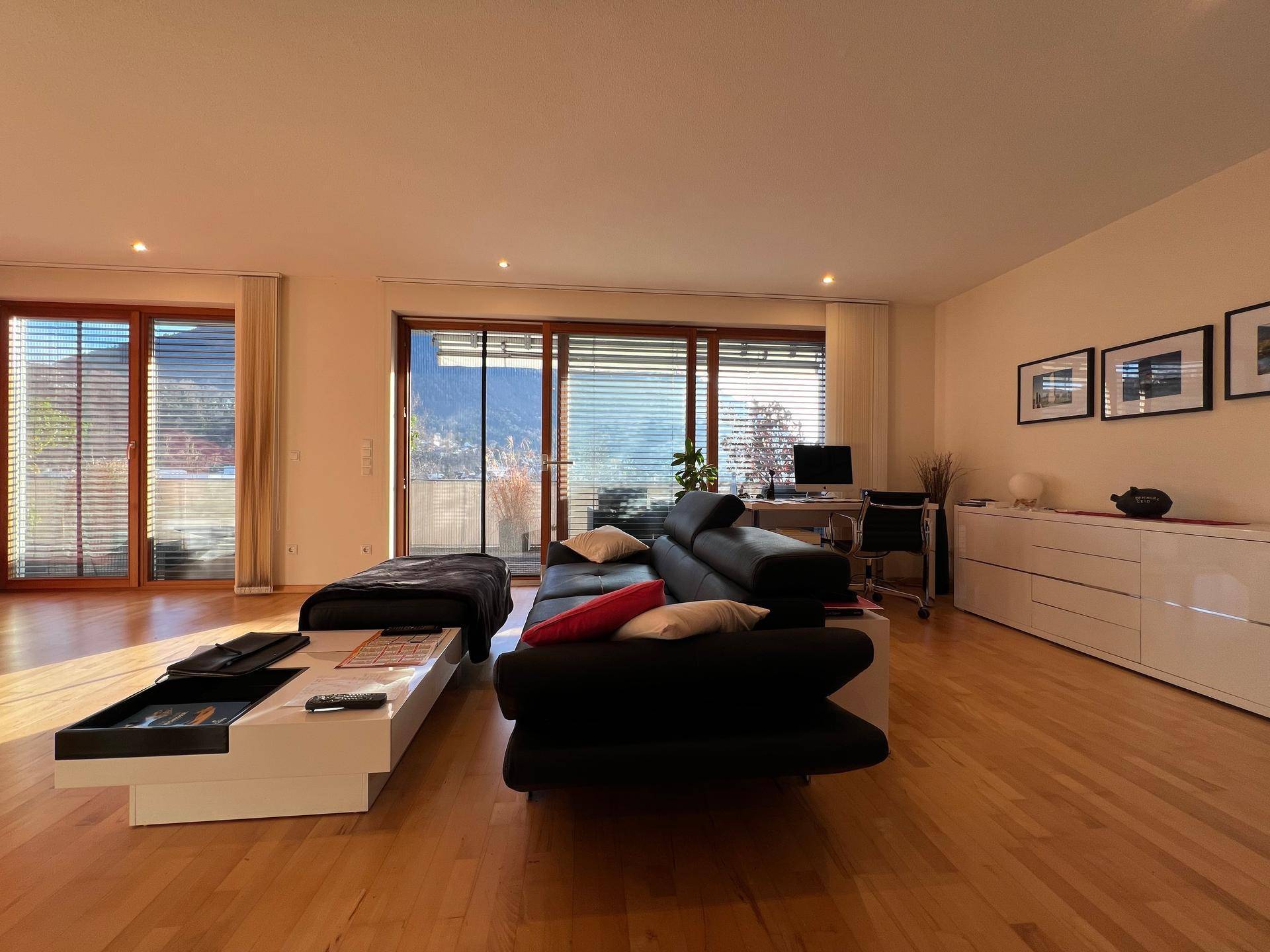 Geräumiges Wohnzimmer mit Holzboden, bequemer Couch und Arbeitsbereich am Fenster.