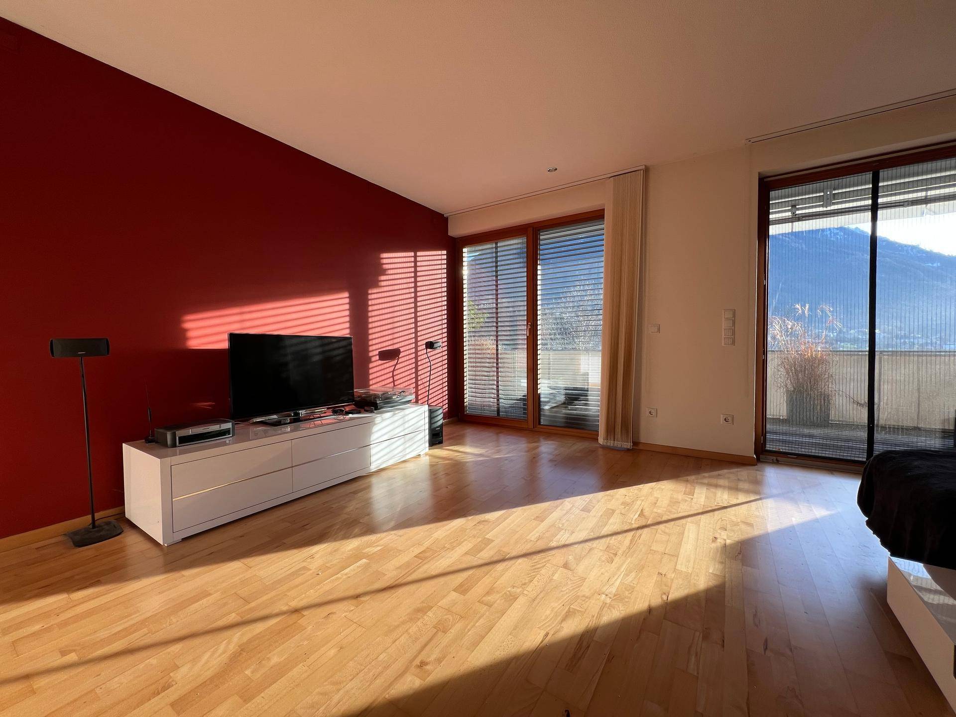 Helles Wohnzimmer mit Holzboden, roter Akzentwand und Zugang zum Balkon mit Bergblick.