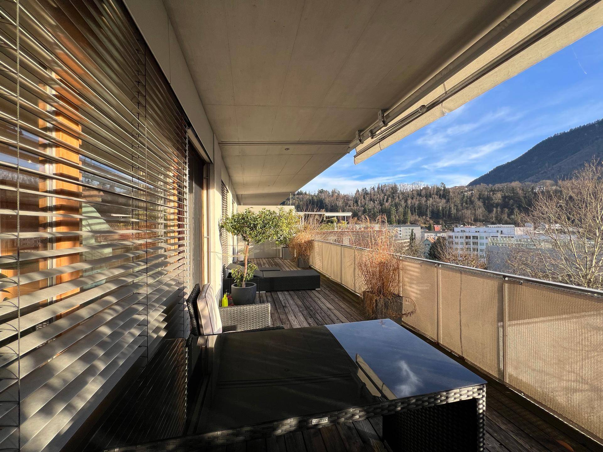 Sonniger Balkon mit Holzboden, Sitzgelegenheiten und weitem Blick auf die Stadt und die Berge.