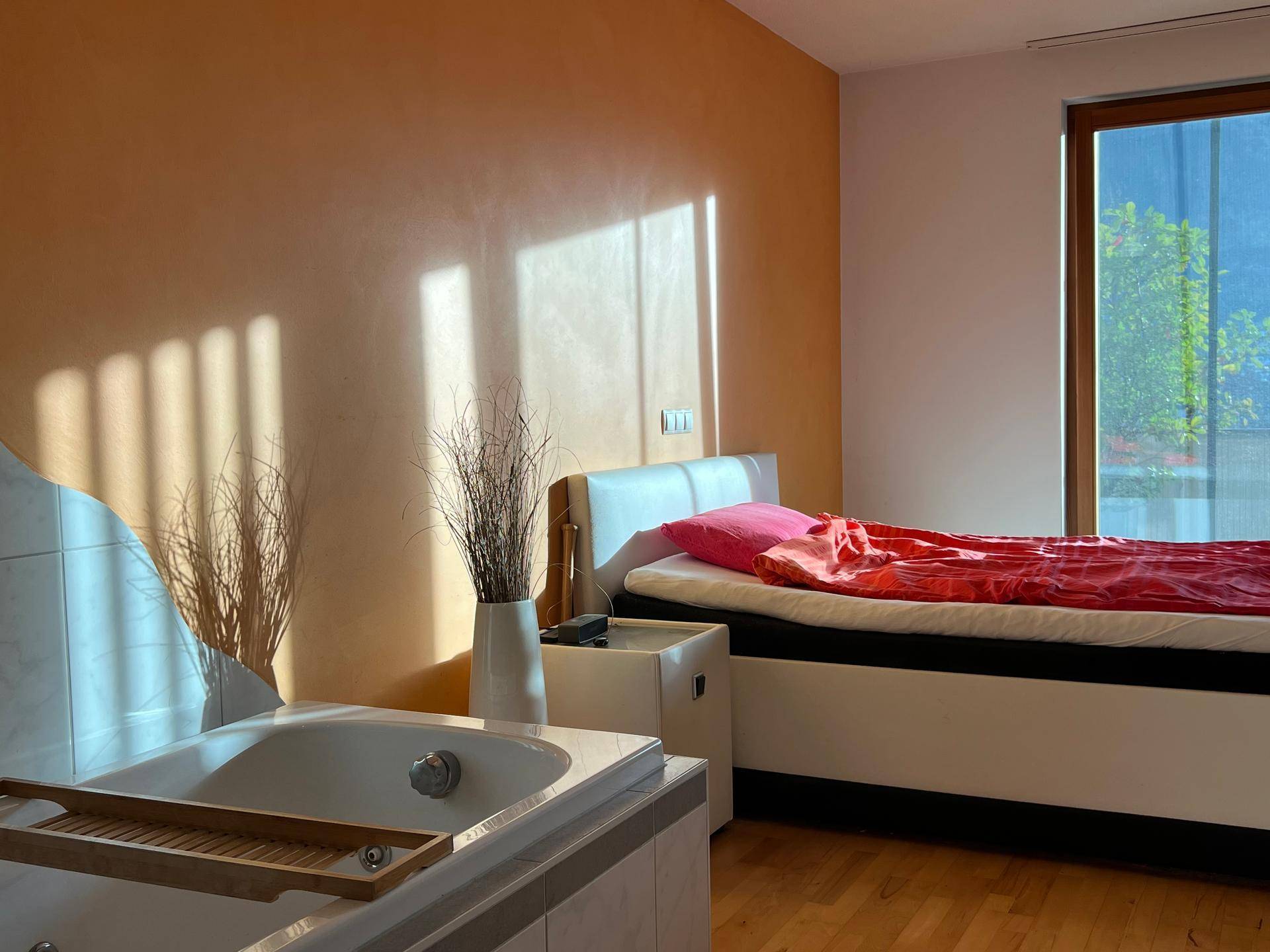 Helles Schlafzimmer mit Holzboden, orangefarbener Wand und integrierter Badewanne.