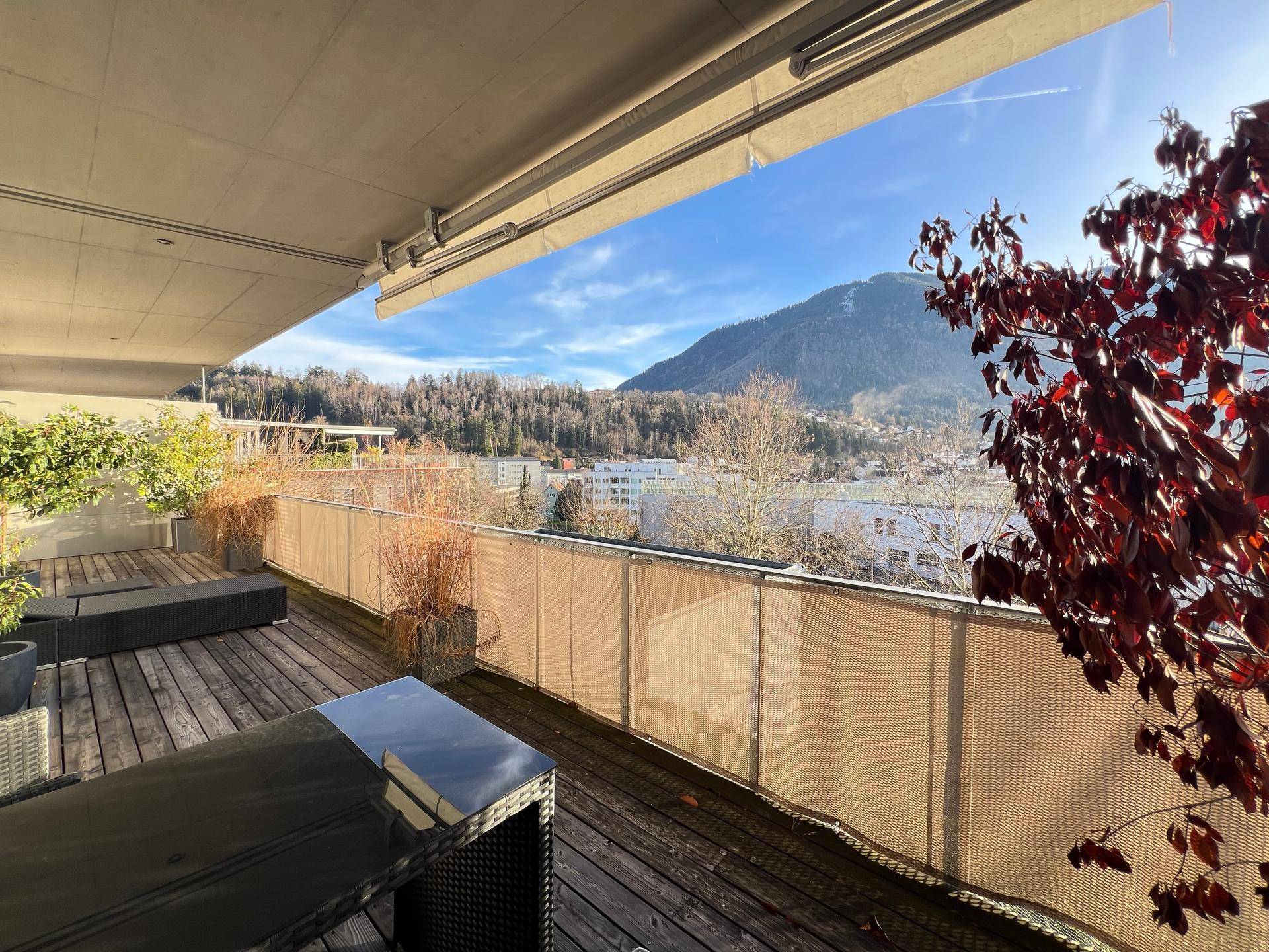 Geräumiger Balkon mit Holzboden, Sitzgelegenheiten und weitem Blick auf die Berge und die Stadt.