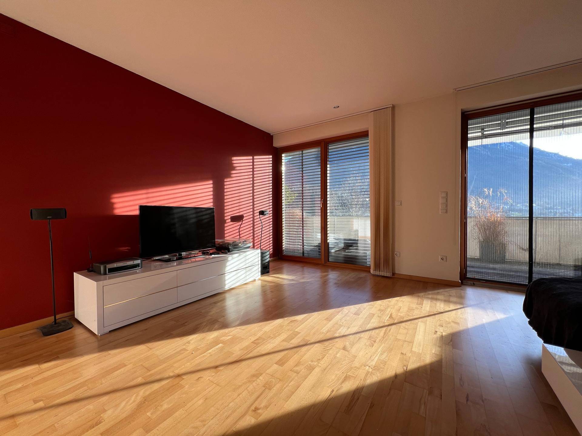 Helles Wohnzimmer mit Holzboden, roter Akzentwand und Zugang zum Balkon mit Bergblick.