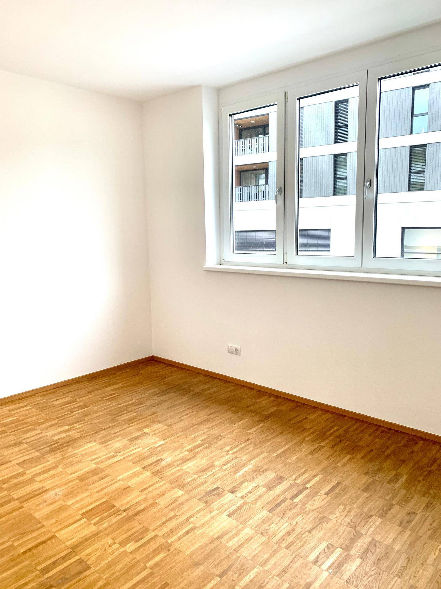Schlafzimmer mit hellem Parkettboden und großem Fenster mit Blick auf die Nachbarbebauung.