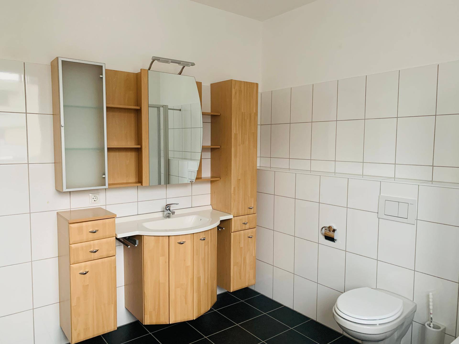 Badezimmer mit weißen Fliesen, dunklem Boden, Holzwaschtisch und Spiegelschrank.
