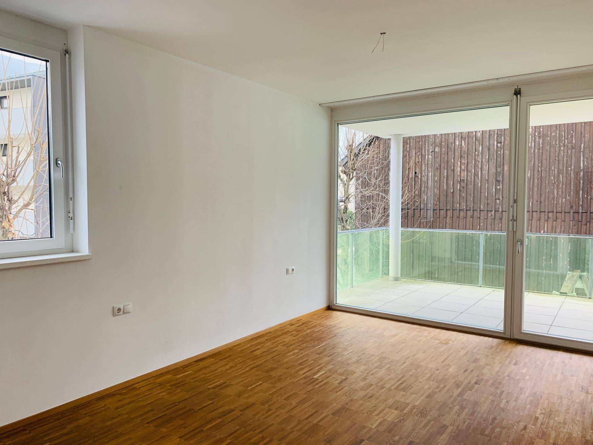Heller Raum mit Parkettboden, großem Fenster und Zugang zu einer Außenterrasse.