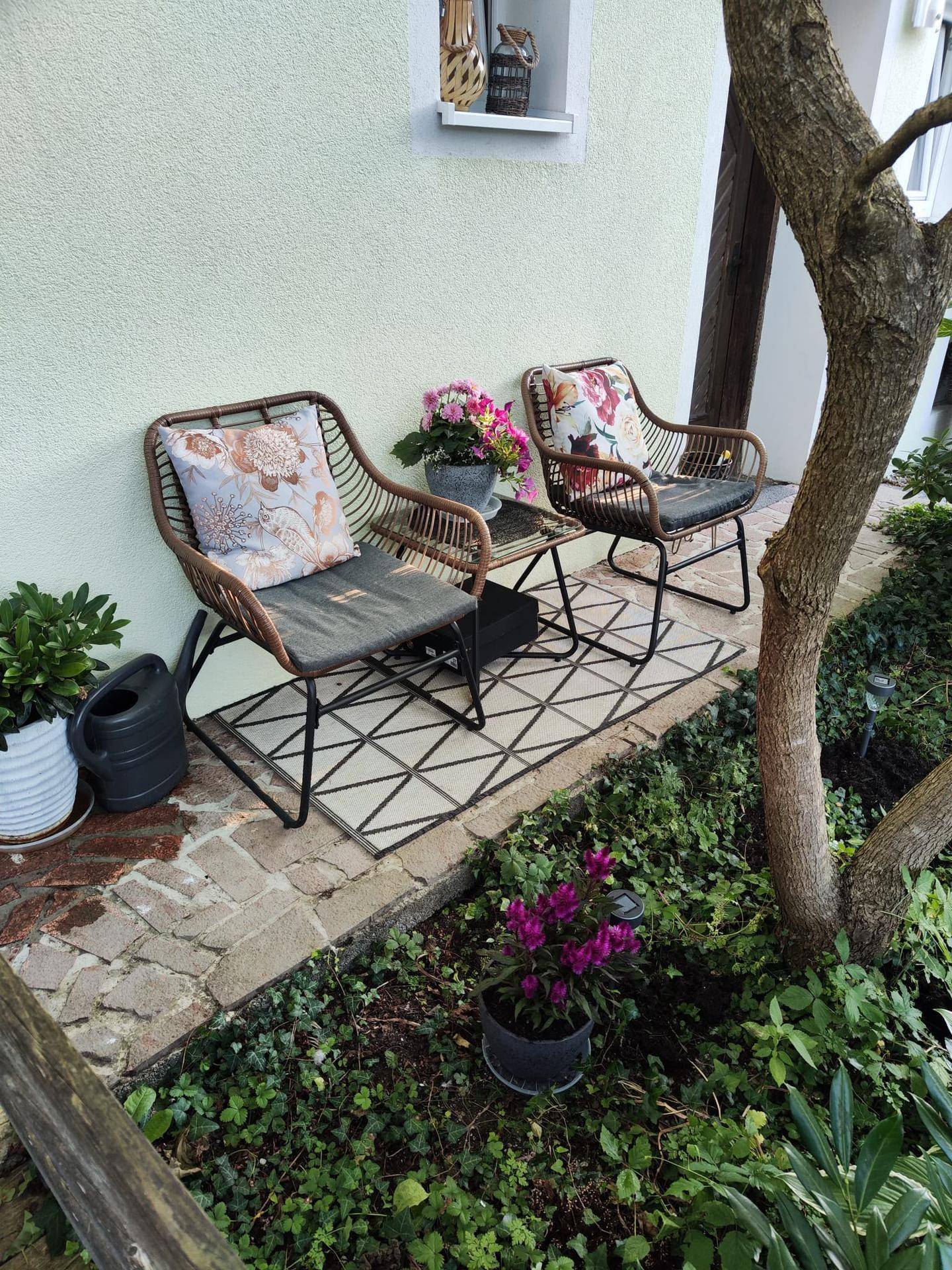 Gemütlicher Sitzbereich im Freien mit Rattanmöbeln und Pflanzen in einer grünen Umgebung.