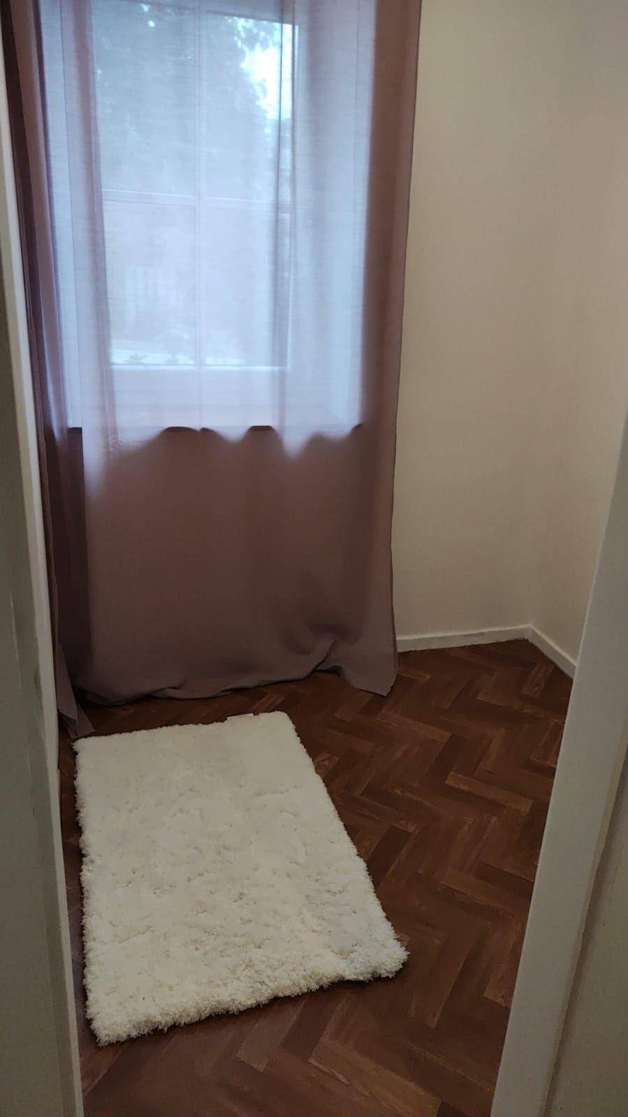 Kleiner Innenraum mit Fenster, Vorhang und einem weißen Teppich auf Parkettboden.