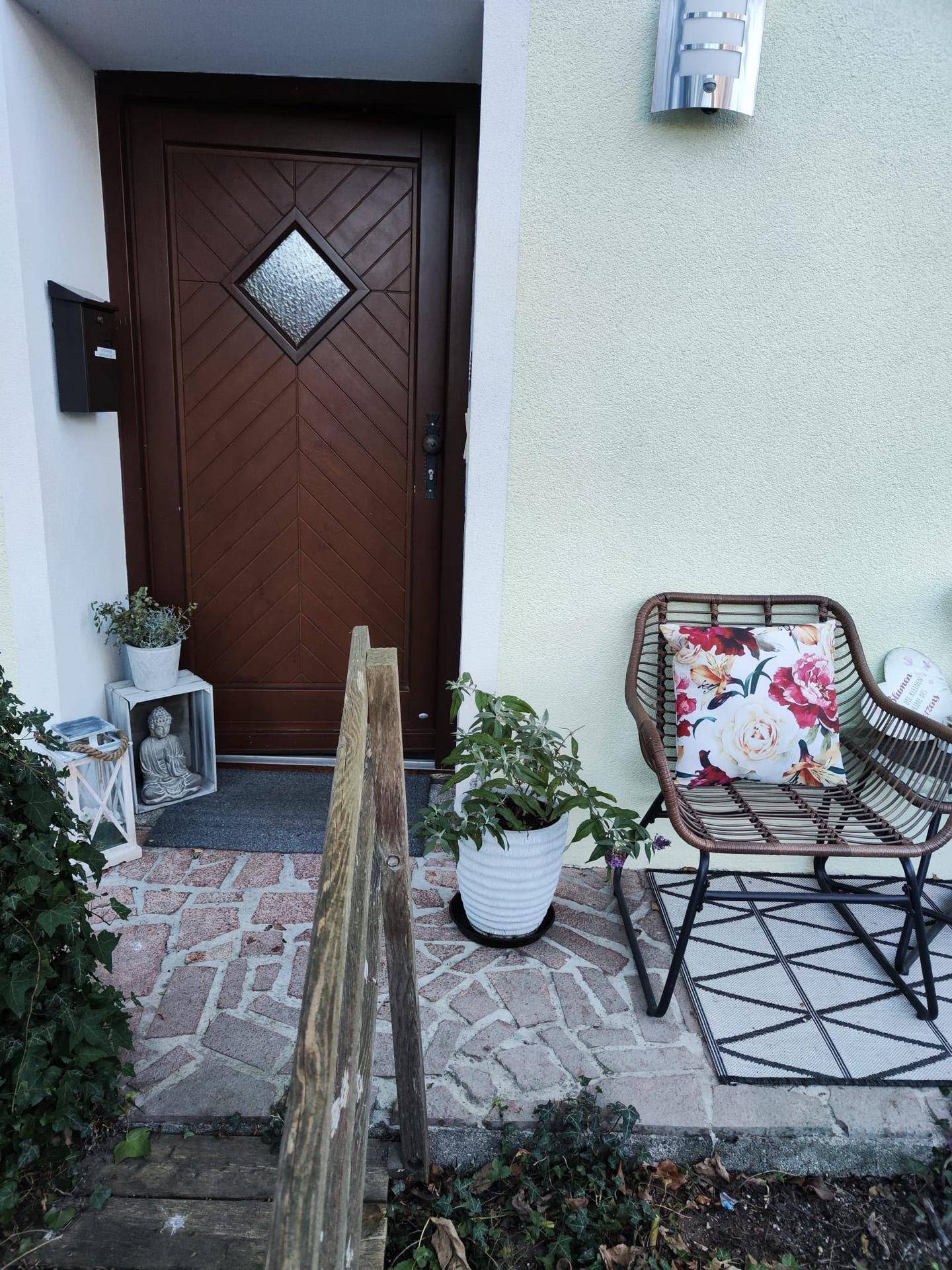 Eingangsbereich des Hauses mit brauner Tür, heller Fassade und dekorativen Pflanzen.