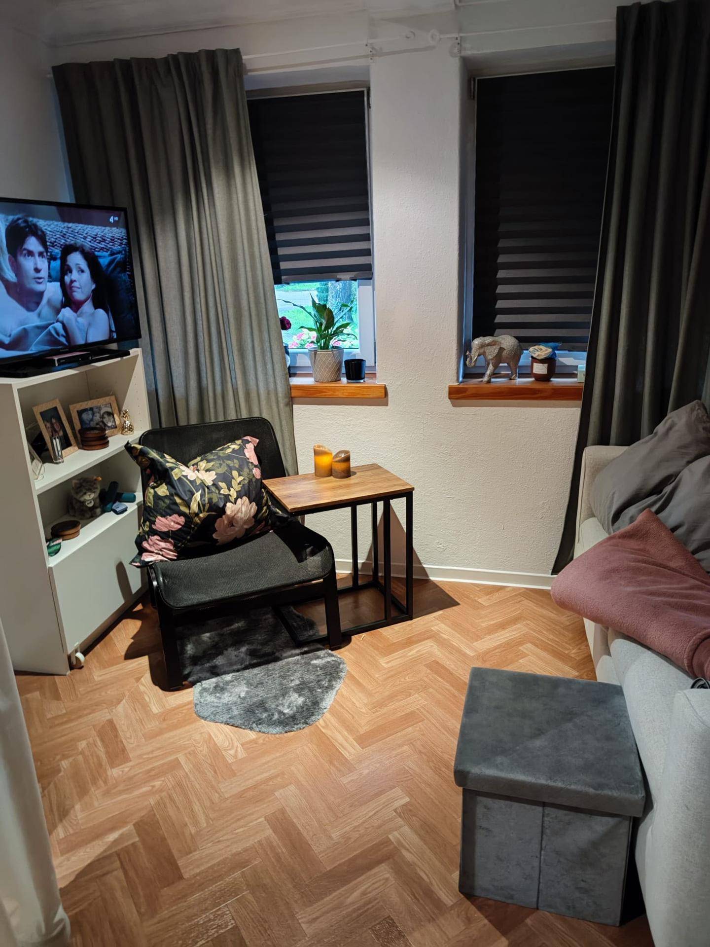 Gemütliches Wohnzimmer mit Fernseher, Sessel und Sofa, ausgestattet mit Vorhängen und Parkettboden.