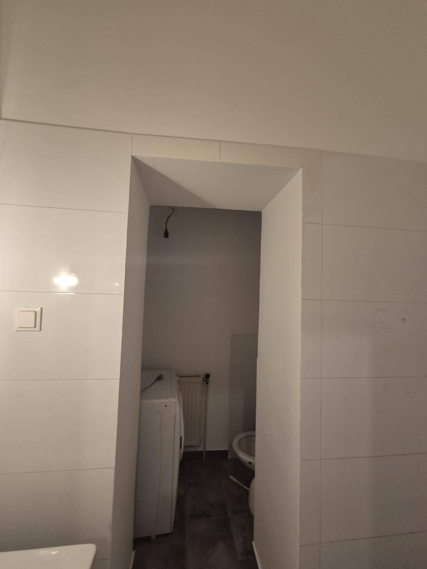 Schmaler, gefliester Raum mit Waschmaschine und Toilette im Hintergrund.