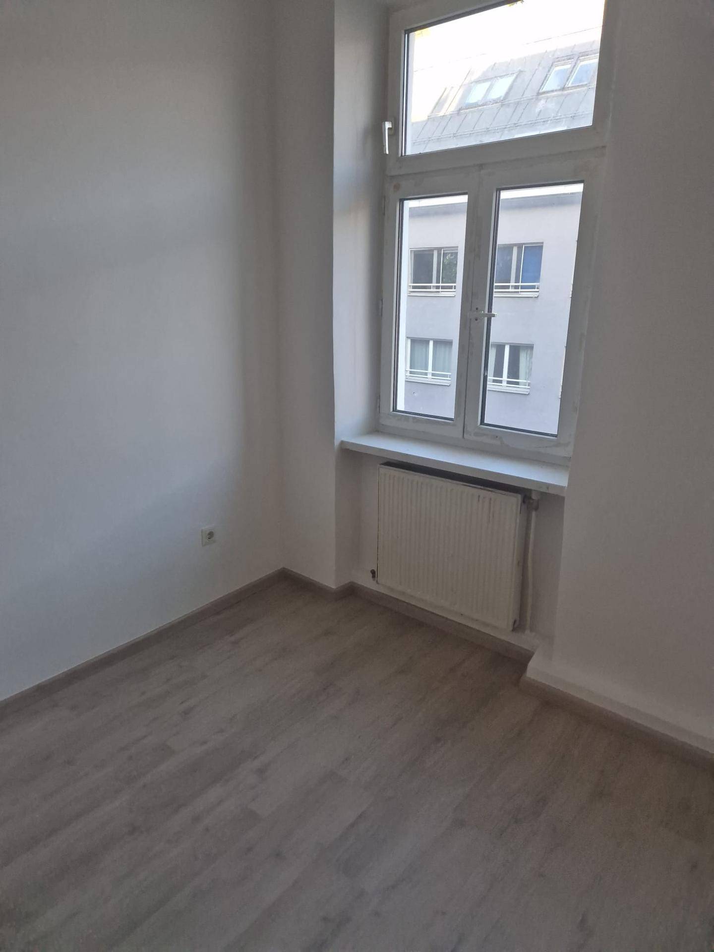 Leeres Zimmer mit hellem Laminatboden, weißen Wänden und Blick auf umliegende Gebäude.