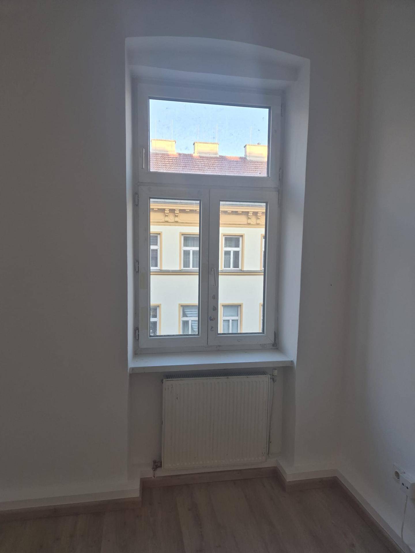 Helles, leeres Zimmer mit Laminatboden, weißen Wänden und Blick auf Dächer und Gebäude.