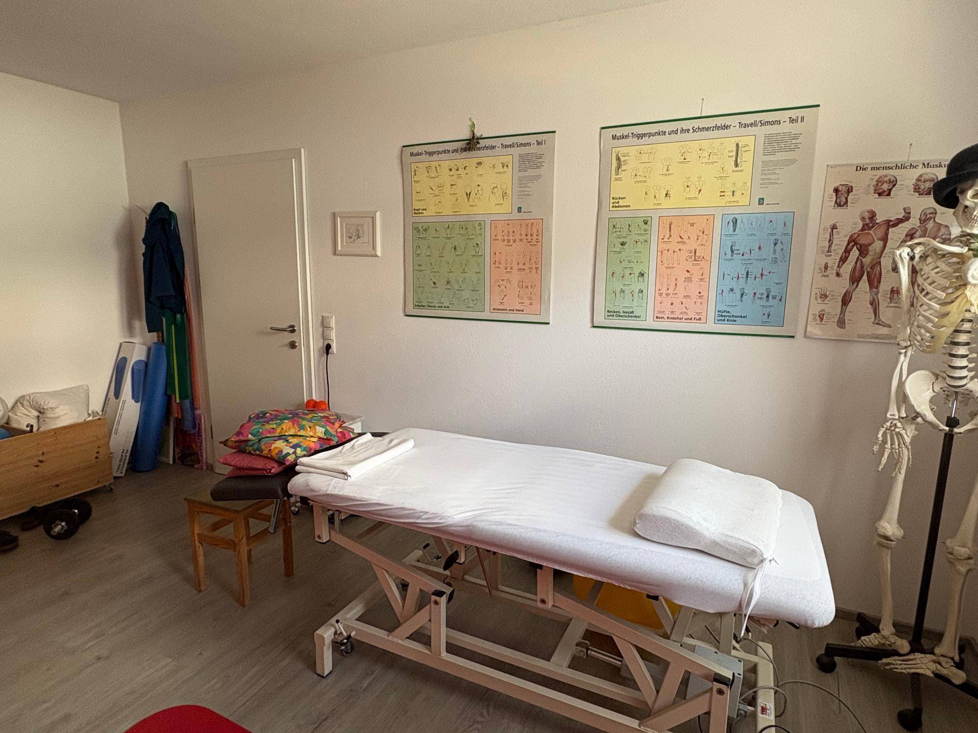 Therapieraum aus einer anderen Perspektive, mit Behandlungsliege, Postern und einem geöffneten Schrank.