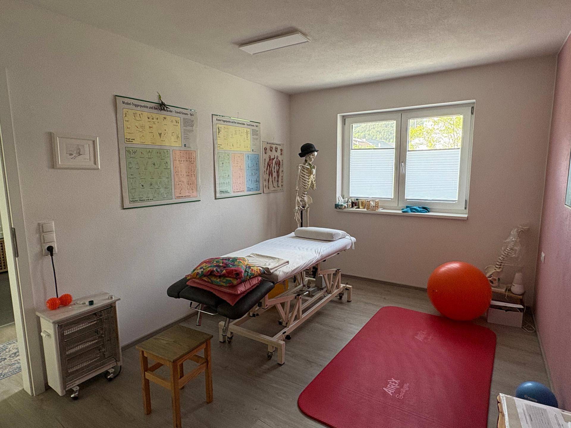Therapieraum mit einer Behandlungsliege, anatomischen Postern, einem Skelettmodell und großen Fenstern.