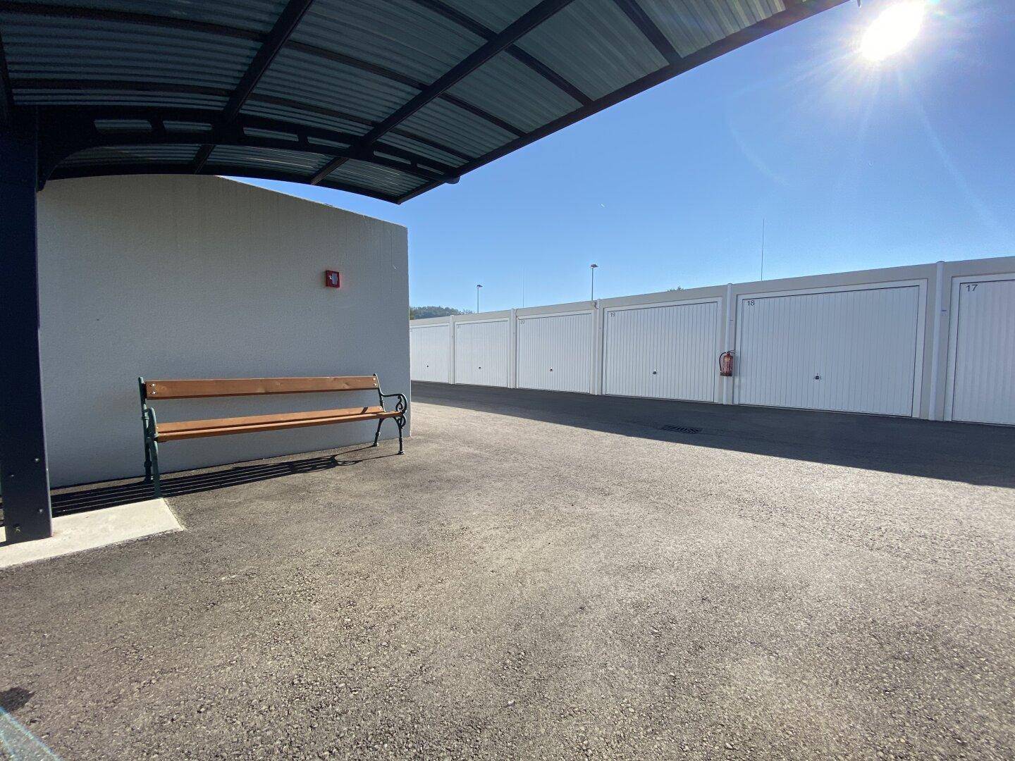 Blick vom überdachten Bereich entlang der Garagenreihe mit Sitzbank und klarem Himmel.