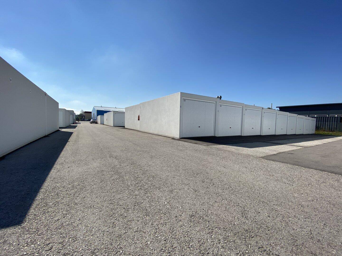 Lange Reihe von weißen Garagenboxen mit industrieller Umgebung im Hintergrund unter klarem Himmel.