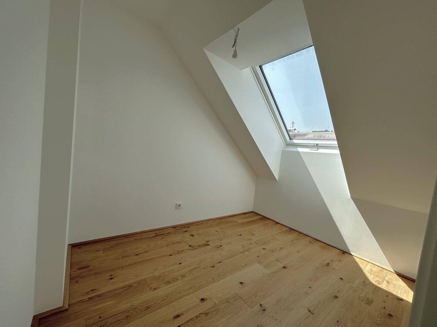 Lichtdurchflutetes Dachzimmer mit Holzboden und schrägem Dachfenster für viel Tageslicht.