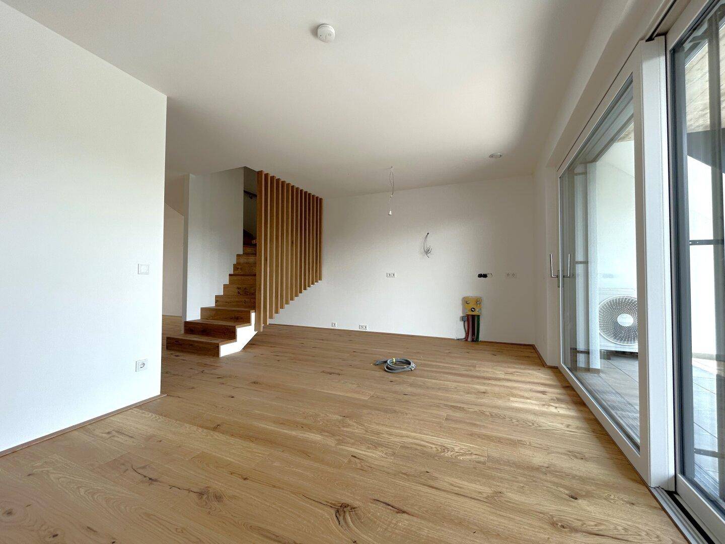 Geräumiger Wohnbereich mit Eichenparkett, Holztreppe und Zugang zu einem Balkon.