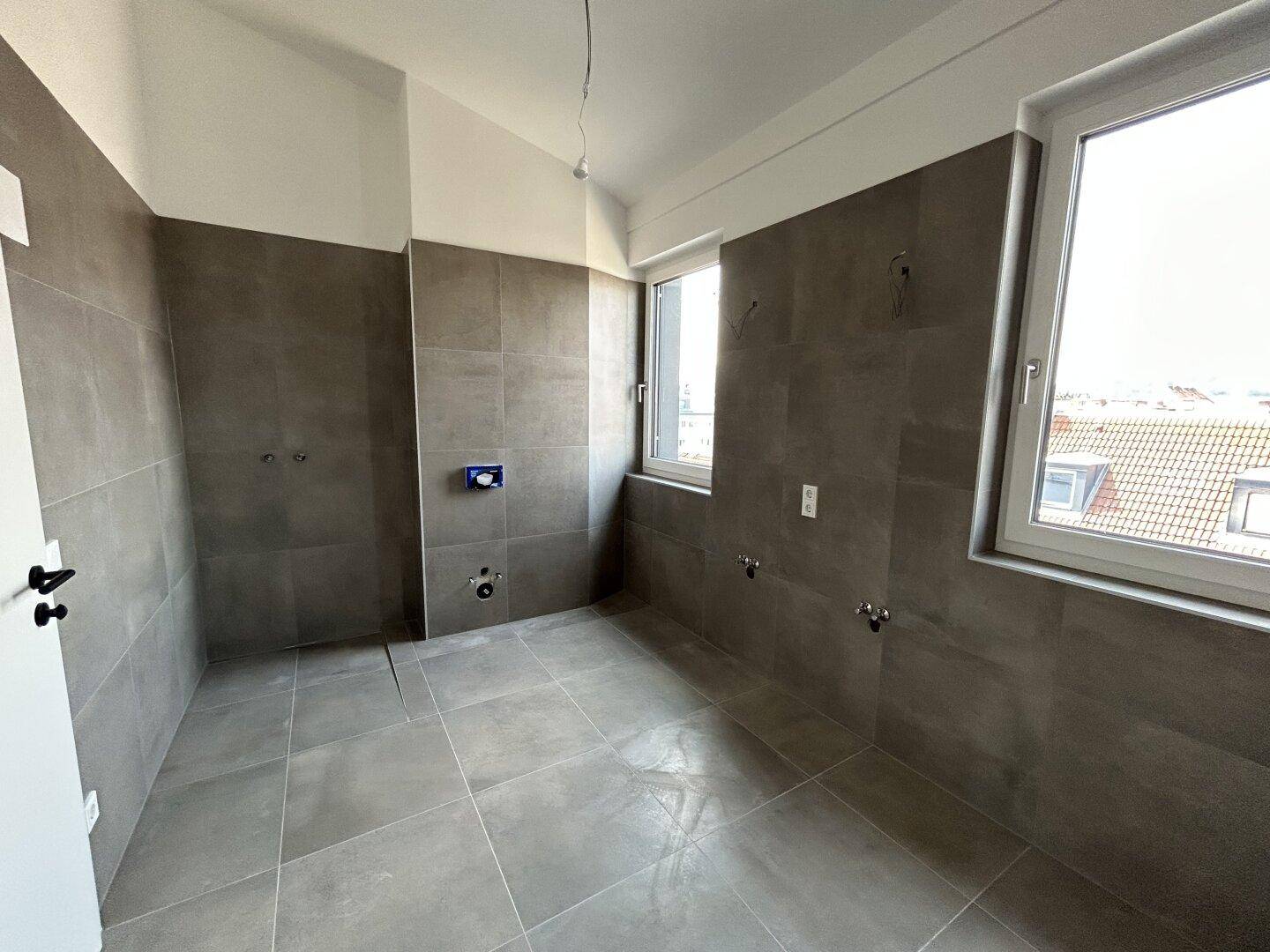 Modernes Badezimmer im Rohbau mit grauen Fliesen und zwei Fenstern.